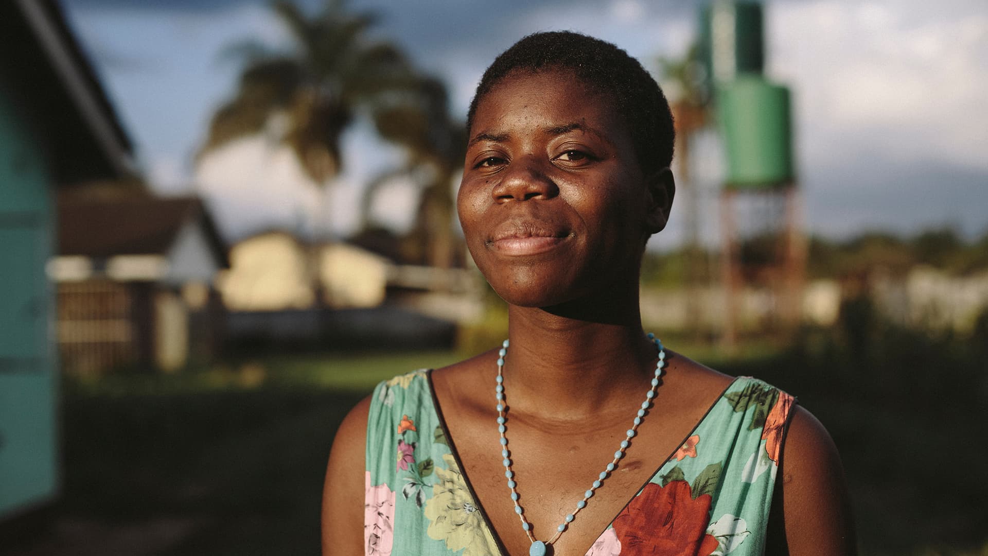Foto: Anna Hugosson Kvinna i Zimbabwe tittar in i kameran. I bakgrunden syns låga byggnader och fält av gräs.