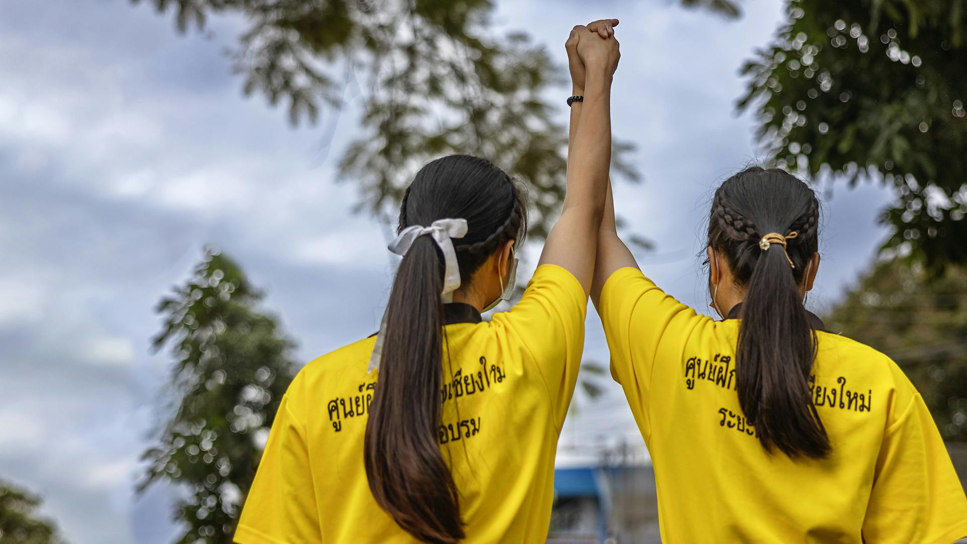 Två unga kvinnor med likadana gula tröjor står med ryggarna mot kameran och håller sina händer upp mot himlen.