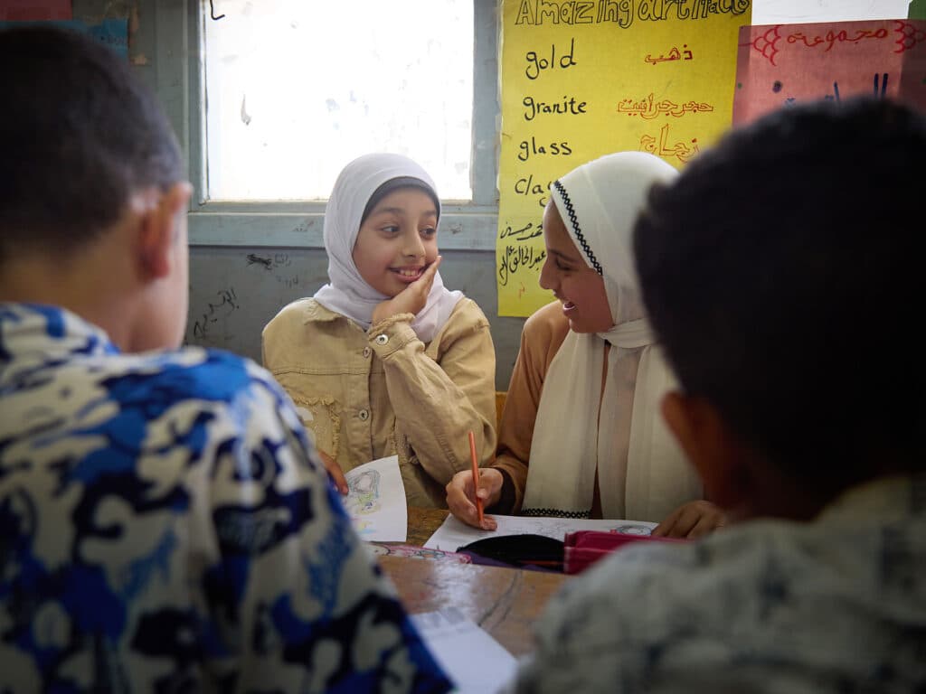 Två flickor i vit hijab sitter i en skolbänk och pratar med varandra.