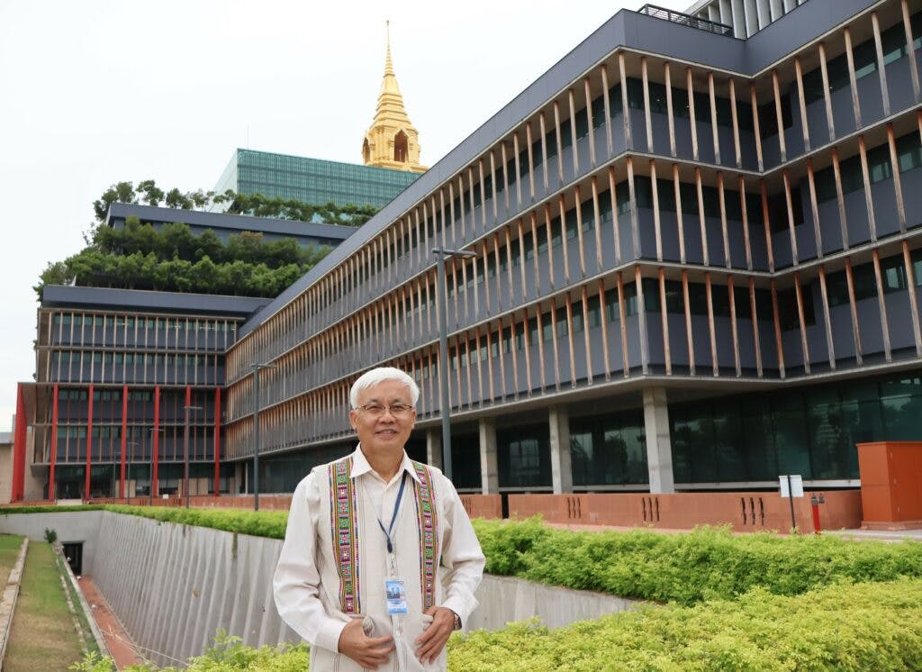 Sakda in a traditional white vest standing in front of a Thai parliament with a golden stupa visible in the background.