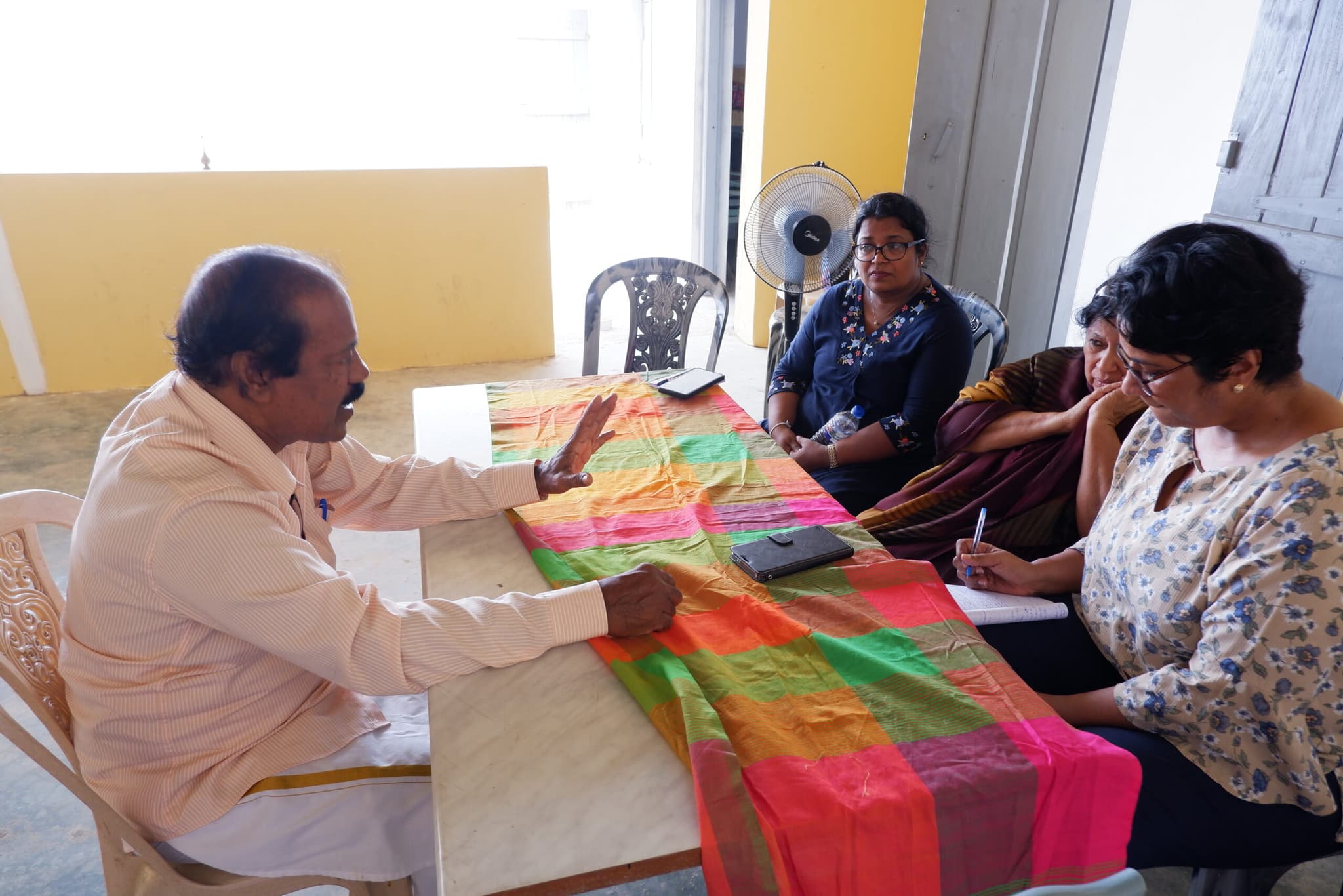 Mr. Chandralingam sitting at a table covered with a colourful cloth, engaged in conversation with Diakonia and partner, MWRAF