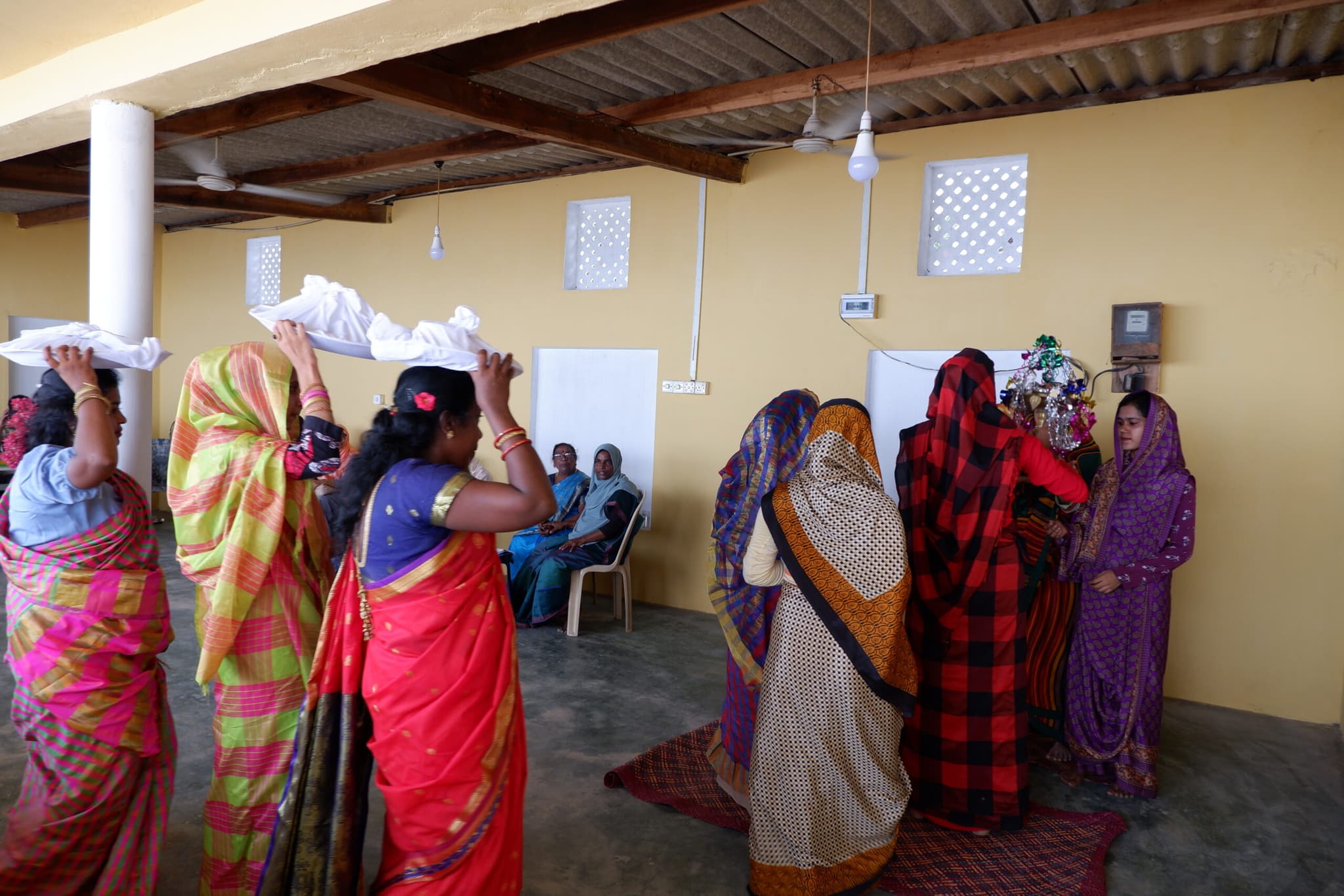 A group of women dressed in vibrant sarees are seen walking in a line, balancing items wrapped in white cloth on their heads. Other women are seated in the background, observing the activity in a room.