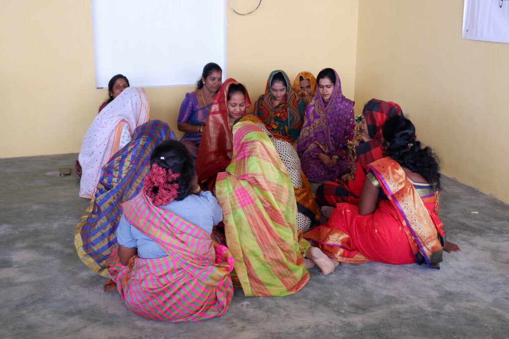 Women are seated on the floor in a circle, draped in colourful clothes, engaged in a group activity.