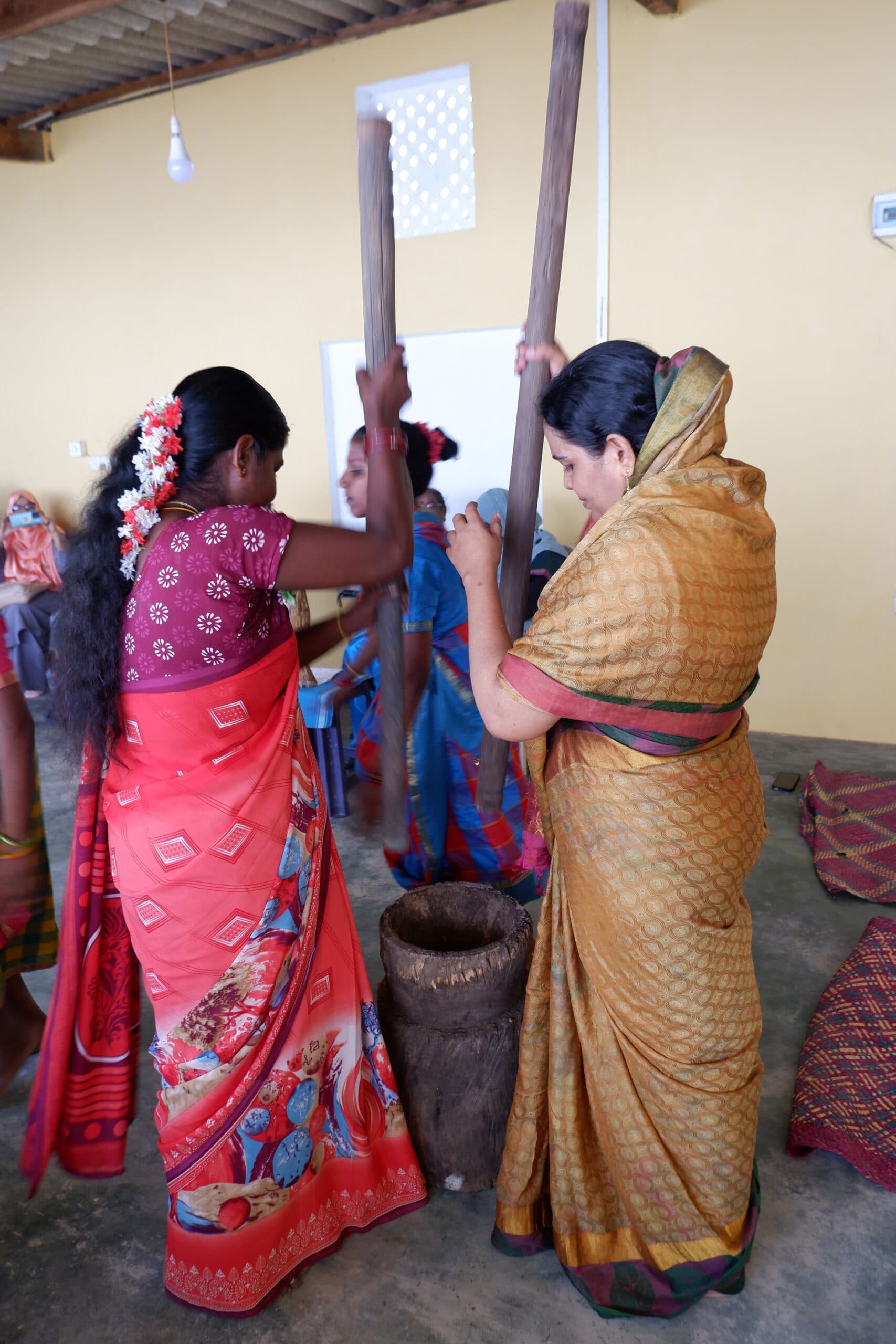 Two women stand side by side, holding large wooden pestles, grinding in a traditional mortar.