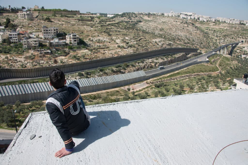 A person on a rooftop overlooking the high concrete wall and settlements on a hill. A person on a rooftop overlooking the high concrete wall and settlements on a hill.