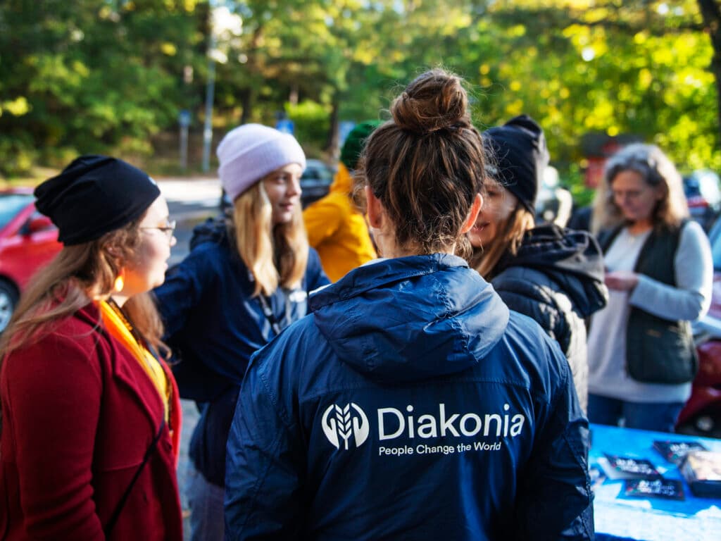 Photo: Diakonia A petrson standing with their back against the camera wearing a Diakonia jacket. In the background there are more people.