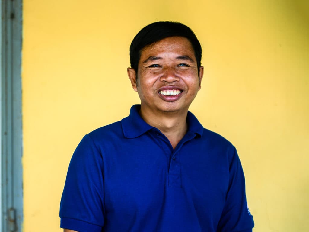 Photo: Chris Cusick A portrait of a smiling man in a blue t-shirt against a yellow wall.
