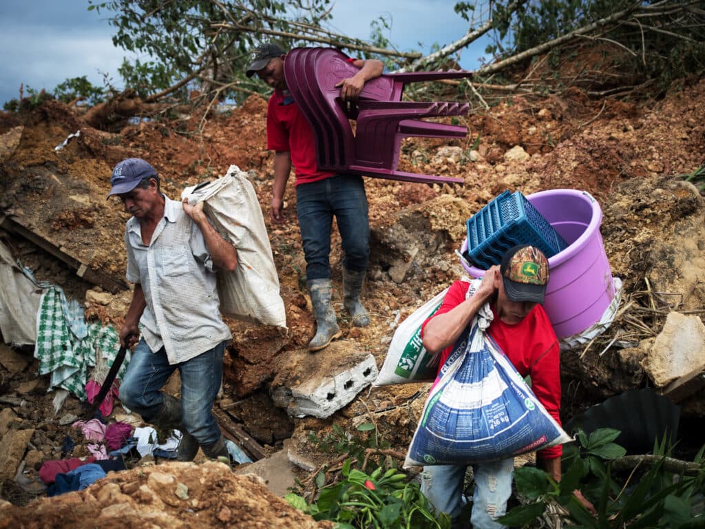 Three men walking down a hill, carrying bags, chairs and other large objects.